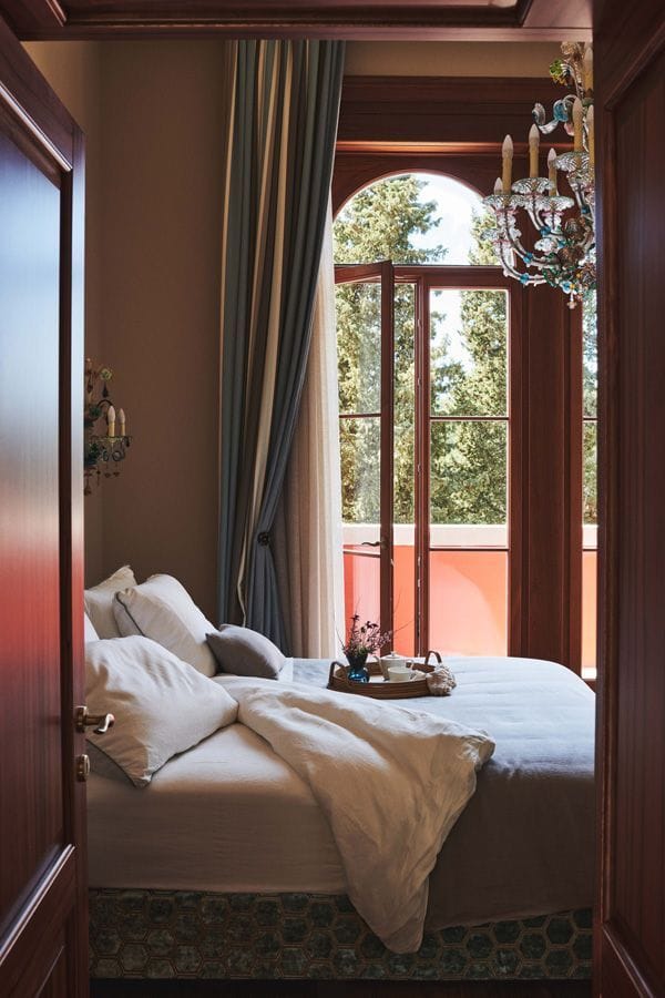 A view of a cozy bedroom with an inviting bed, elegant drapes, a colorful chandelier, and a sunny window overlooking trees